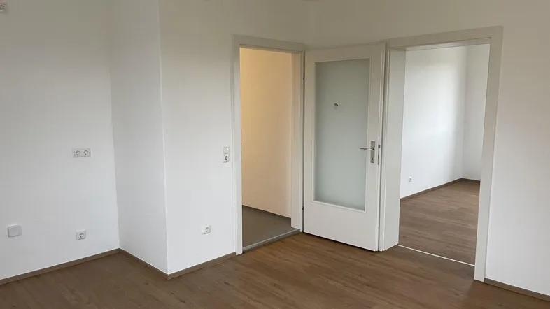 Helles Zimmer mit Laminatboden und zwei Türen, die zu weiteren Räumen führen.
