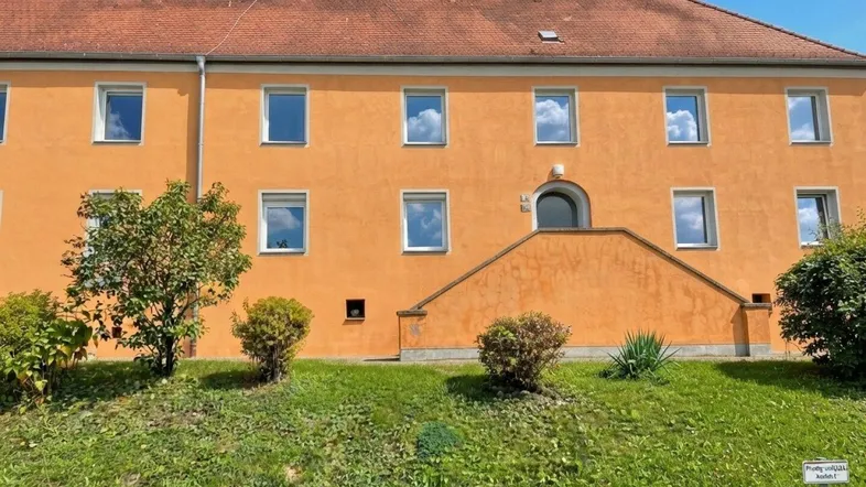 Gebäude mit leuchtend oranger Fassade, rotem Ziegeldach und grünem Vorgartenbereich.