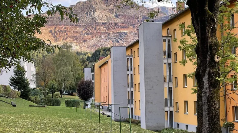 Mehrere Wohngebäude mit gelben und orangen Fassaden vor einer beeindruckenden Bergkulisse und grüner Wiese.