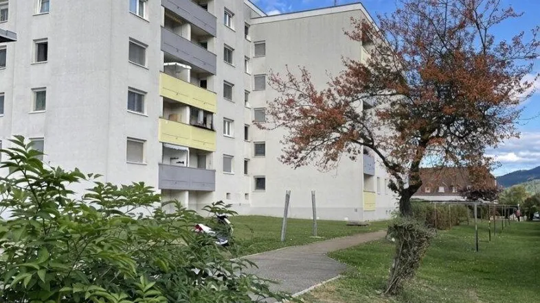 Mehrfamilienhaus mit mehreren Etagen und Balkonen in einer grünen Umgebung unter bewölktem Himmel.