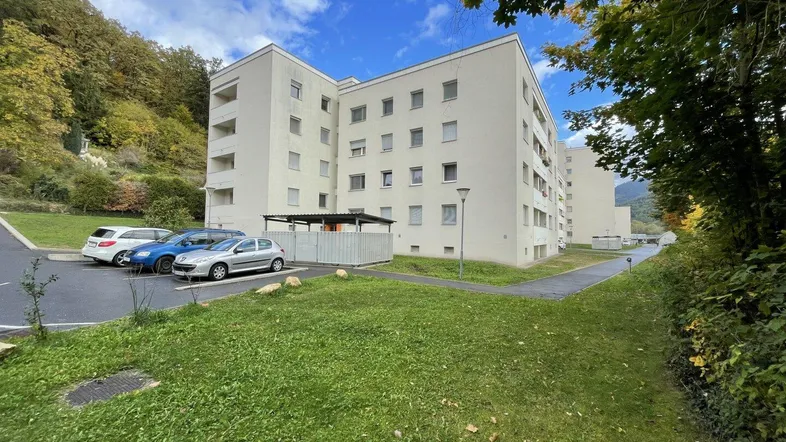Mehrfamilienhaus mit Parkplätzen und grüner Umgebung am Fuße eines bewaldeten Hügels.