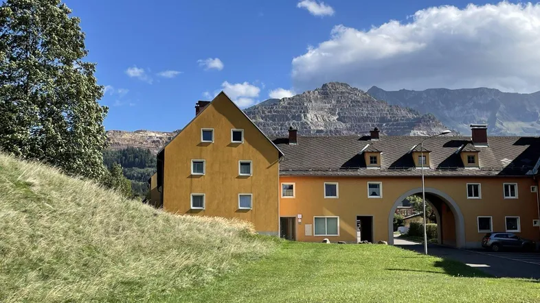 Gebäude mit gelber Fassade und dunklem Dach vor einer beeindruckenden Bergkulisse mit einem Steinbruch.
