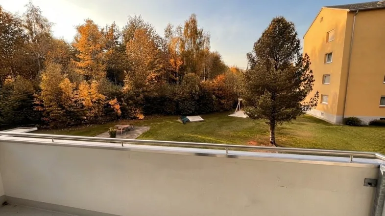 Blick vom Balkon auf eine grüne Fläche mit Bäumen in Herbstfarben und ein angrenzendes Gebäude.