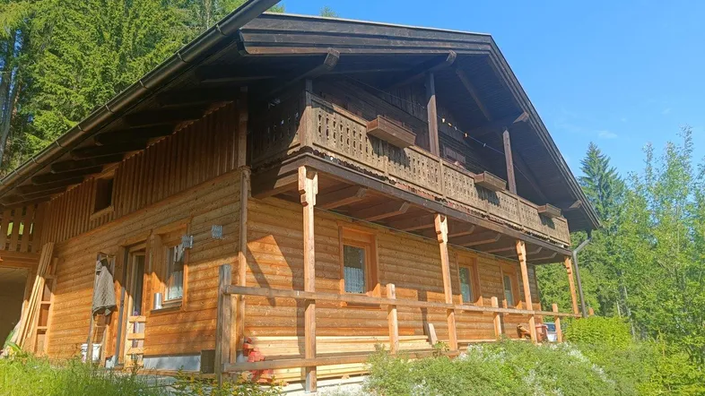 Traditionelles Holzchalet in alpiner Umgebung mit Balkonen und viel Grün unter blauem Himmel.