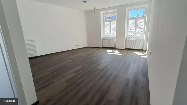 Helles, leeres Wohnzimmer mit modernem Laminatboden und zwei großen Fenstern.