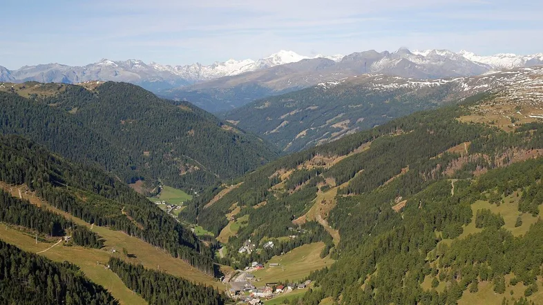 Panoramablick auf ein weites Alpental mit bewaldeten Hängen und schneebedeckten Berggipfeln in der Ferne.