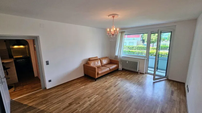 Helles Wohnzimmer mit Parkettboden, Ledersofa und Zugang zu einem Balkon mit Blick ins Grüne.
