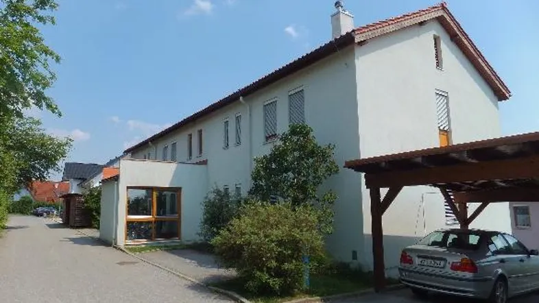 Mehrfamilienhaus mit weißer Fassade, Satteldach und überdachtem Stellplatz für ein Fahrzeug.