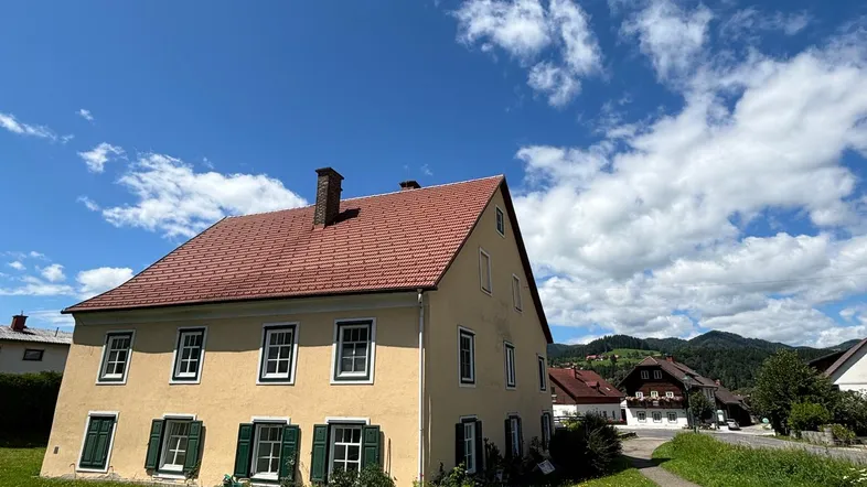 Traditionelles gelbes Haus mit rotem Ziegeldach und grünen Fensterläden unter blauem Himmel.