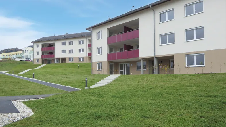 Moderne Wohngebäude mit hellen Fassaden, roten Balkonen und gepflegten Grünflächen in ländlicher Umgebung.