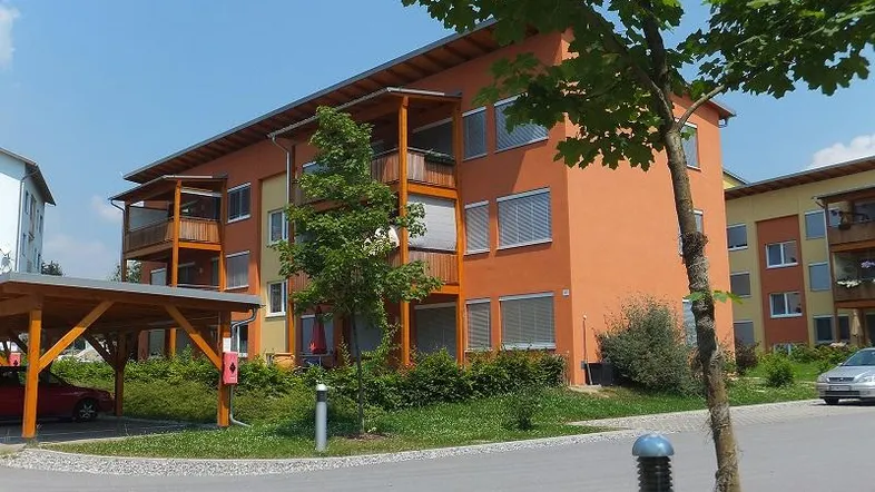 Modernes Mehrfamilienhaus mit orangefarbener Fassade, Balkonen und einem Carport im Grünen.