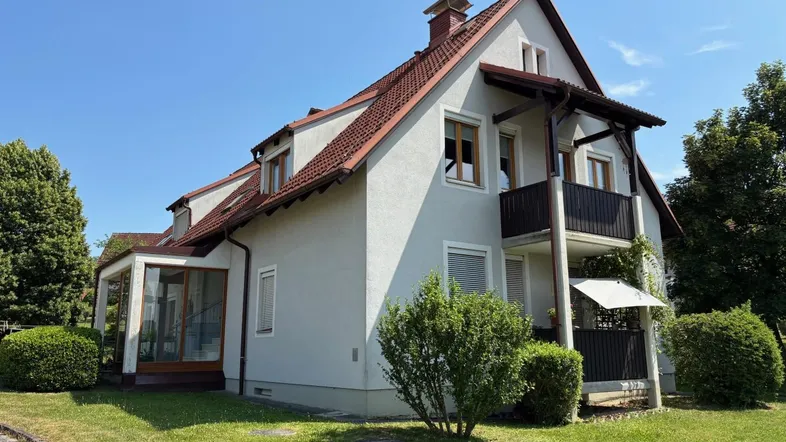 Gepflegtes Haus mit Satteldach, Balkon, Wintergarten und umgebendem Grünbereich unter blauem Himmel.