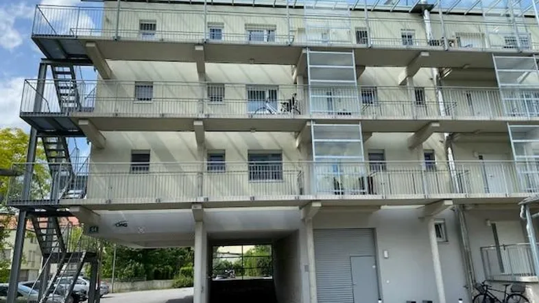 Modernes Mehrfamilienhaus mit mehreren Balkonen, Außentreppe und Tiefgaragenzufahrt, umgeben von Grünflächen.
