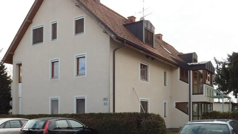 Mehrfamilienhaus mit Satteldach, beiger Fassade und Außenjalousien, umgeben von Hecken und Parkplätzen.