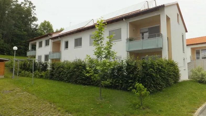 Weißes Mehrfamilienhaus mit Balkonen, grüner Hecke und Solarpaneelen auf dem Dach.