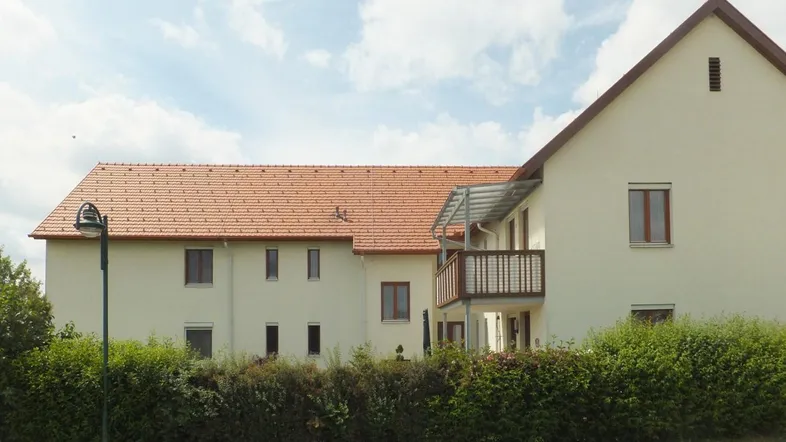 Ansicht eines Mehrfamilienhauses mit heller Fassade, Satteldach und einem Balkon.
