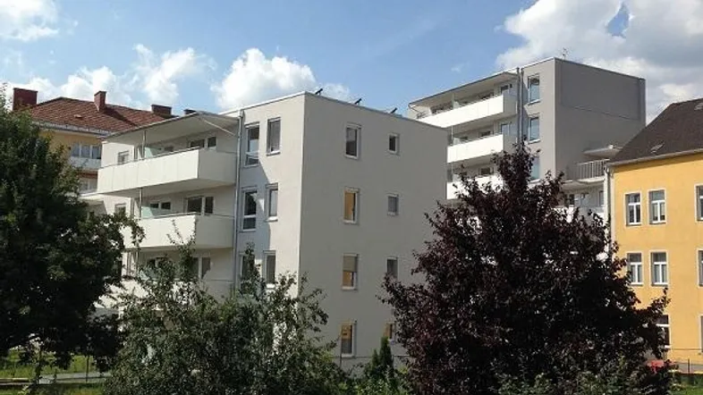 Modernes Mehrfamilienhaus mit hellgrauer Fassade, mehreren Balkonen und umgebender Grünfläche unter blauem Himmel.