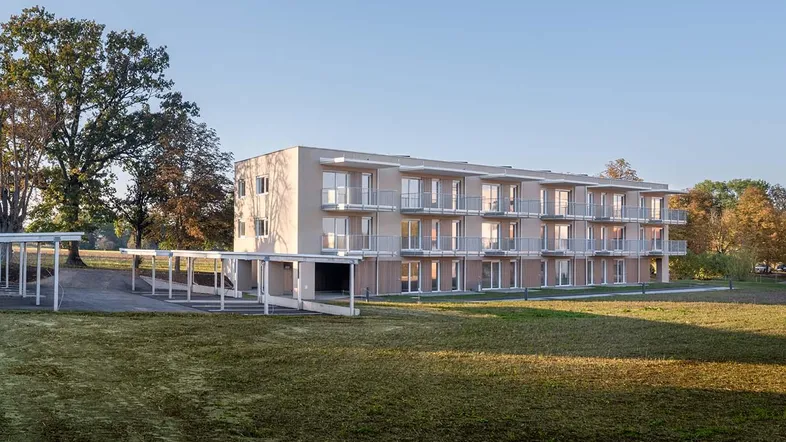 Modernes Mehrfamilienhaus mit zahlreichen Balkonen, umgeben von Grünflächen und mit überdachten Parkplätzen.