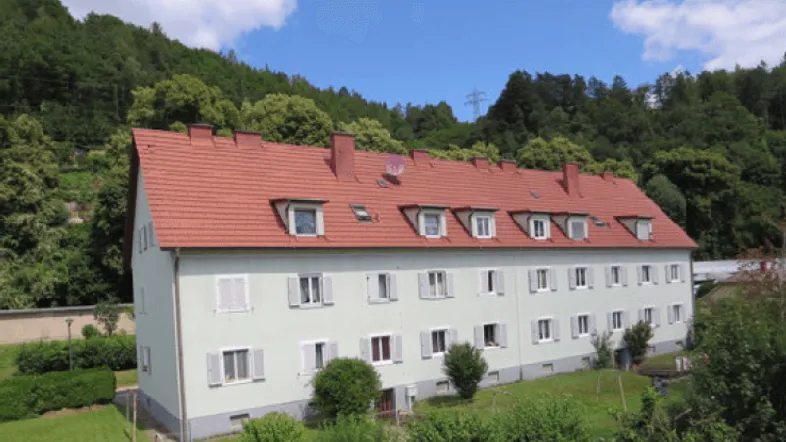 Mehrfamilienhaus mit rotem Mansarddach und hellgrüner Fassade, umgeben von grünen Hügeln und Bäumen.