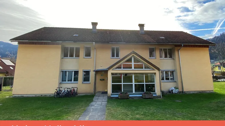 Gelbes Mehrfamilienhaus mit Satteldach, Rasenfläche und verglastem Eingangsbereich.