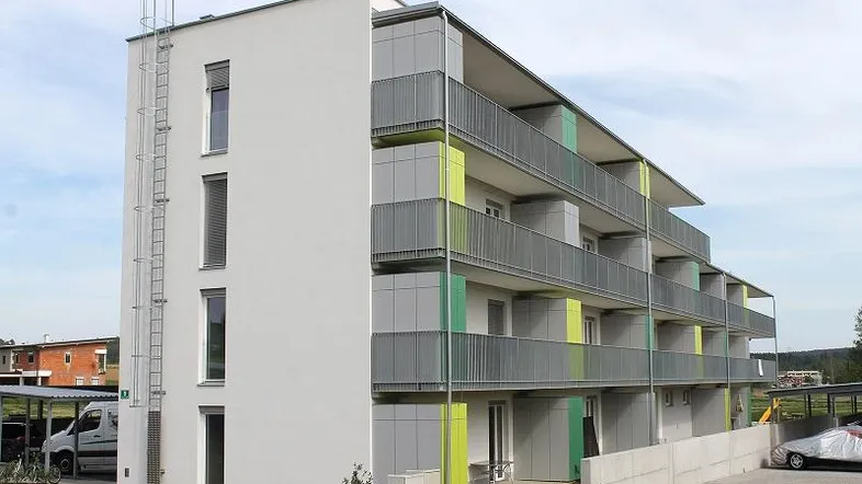 Modernes Mehrfamilienhaus mit Balkonen und farbigen Akzenten an der Fassade, umgeben von Grünflächen.