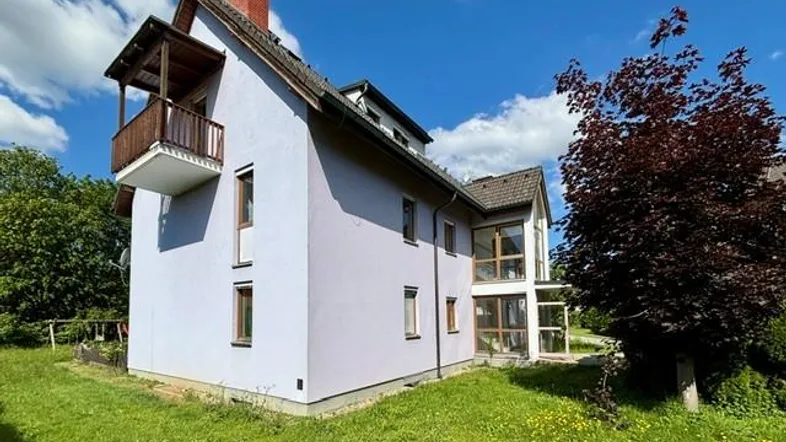 Freistehendes Einfamilienhaus mit hellvioletter Fassade, Holzbalkon und großem Gartenbereich unter blauem Himmel.