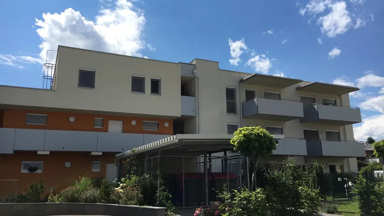 Mehrfamilienhaus mit moderner Fassade, Balkonen und begrüntem Außenbereich unter blauem Himmel.
