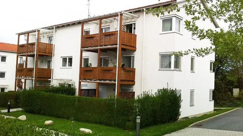 Weißes Mehrfamilienhaus mit Holzbalustraden an den Balkonen und gepflegter Grünanlage.