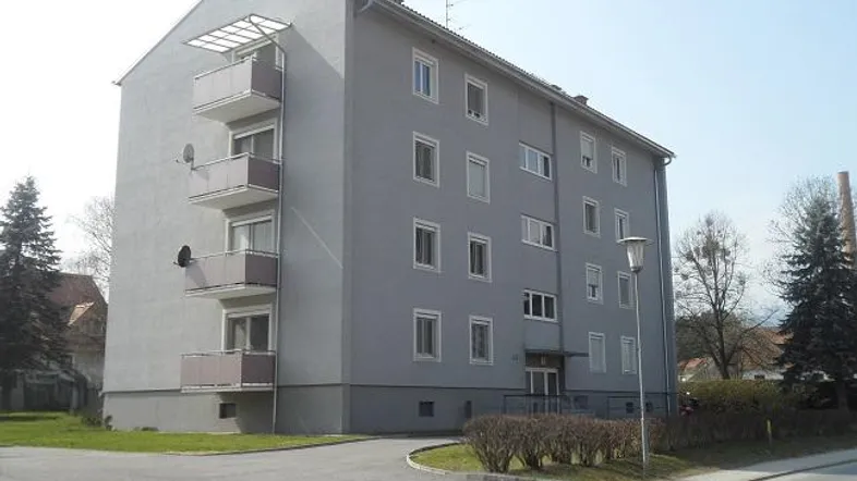Graues Mehrfamilienhaus mit mehreren Balkonen und umliegender Grünfläche unter blauem Himmel.