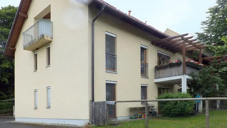 Mehrfamilienhaus mit beiger Fassade, Balkonen, Pergola und einem kleinen Gartenbereich.