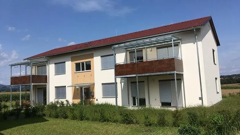 Modernes Mehrfamilienhaus mit Balkonen und rotem Dach in ländlicher Umgebung.