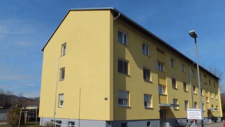 Gelbes Mehrfamilienhaus mit mehreren Etagen und blauer Sockelzone unter klarem Himmel.