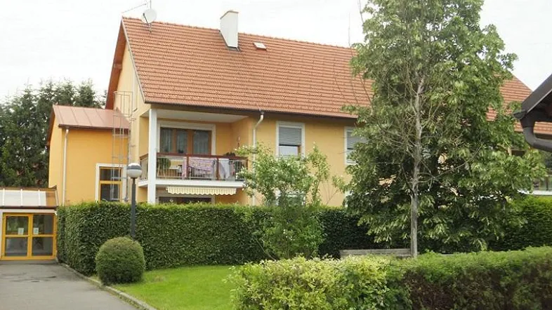 Gelbes Mehrfamilienhaus mit rotem Ziegeldach, Balkon und gepflegter Hecke im Vorgarten.