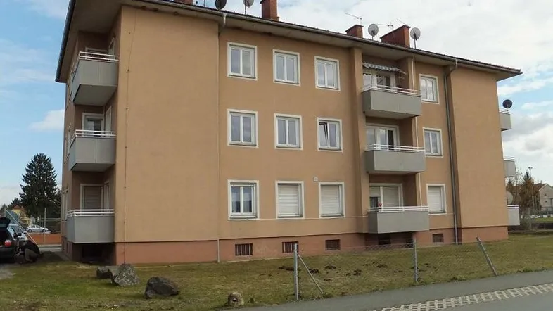 Mehrfamilienhaus mit beigefarbener Fassade, mehreren Balkonen und Satteldach in grüner Umgebung.
