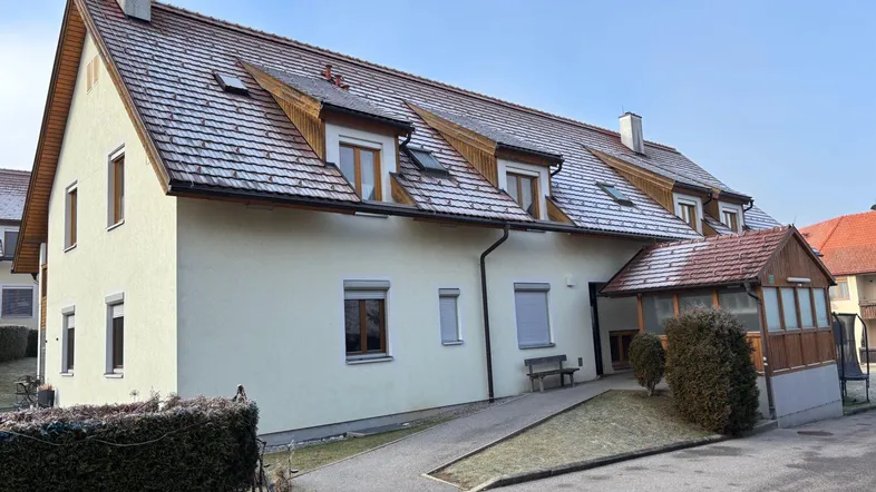 Mehrfamilienhaus mit Satteldach und Gauben, umgeben von einer Hecke und einem Gehweg.