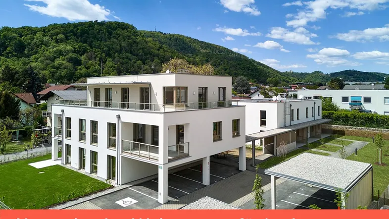 Modernes Mehrfamilienhaus mit Balkonen, Carports und Grünflächen in bergiger Umgebung unter blauem Himmel.