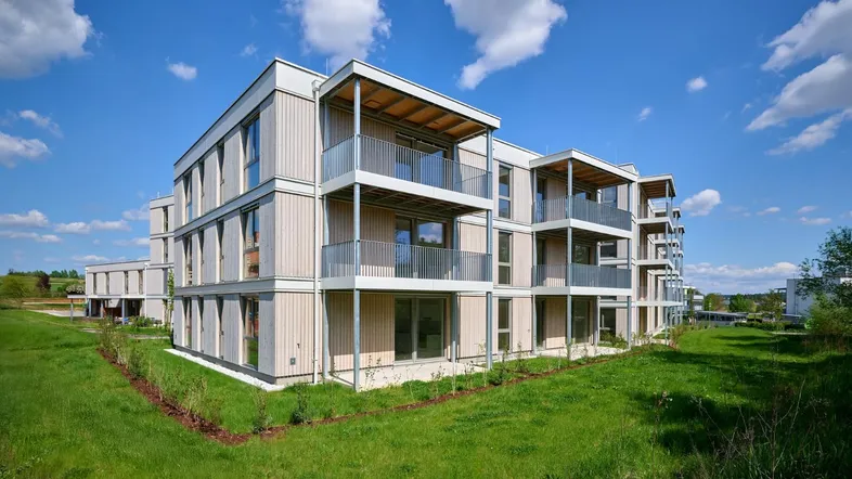 Modernes Mehrfamilienhaus mit hellen Fassaden und Balkonen, umgeben von grüner Landschaft unter blauem Himmel.