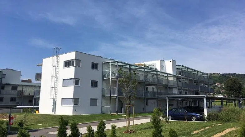 Modernes Mehrfamilienhaus mit Balkonen, Carports und Grünflächen unter blauem Himmel.
