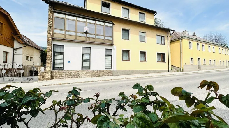 Gelbes Mehrfamilienhaus mit modernem Anbau, Balkon und Rollläden an der Straßenseite.