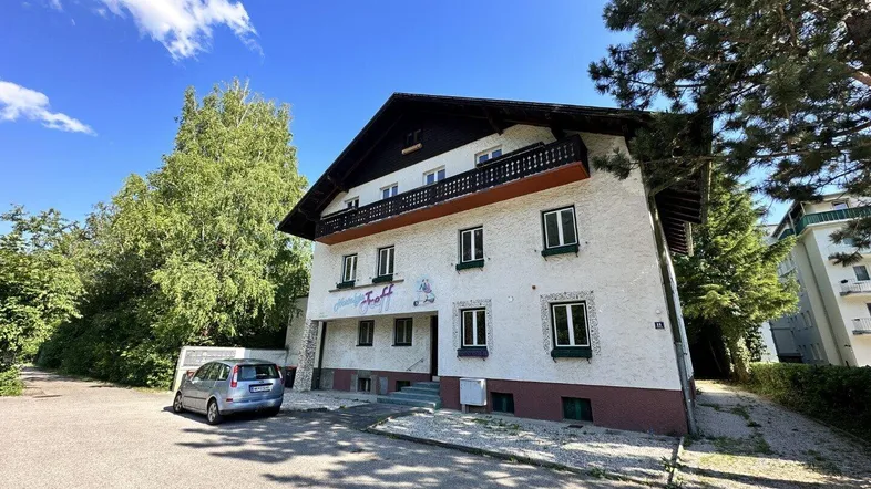 Traditionelles Gebäude mit Satteldach und Balkon, umgeben von Bäumen und mit Parkplätzen.