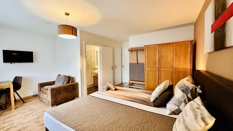 Geräumiges Schlafzimmer mit Doppelbett, Sessel und Holzschrank, angrenzend an ein Badezimmer.