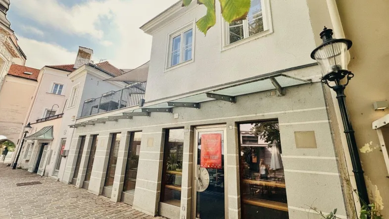 Fassade eines Gebäudes mit Ladenlokalen im Erdgeschoss und einer Terrasse im Obergeschoss an einer belebten Straße.