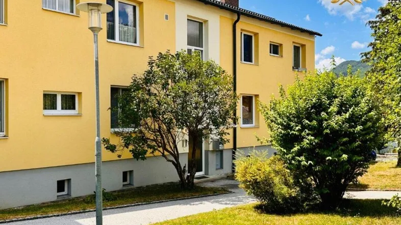 Gelbes Mehrfamilienhaus mit gepflegtem Vorgarten und Bäumen unter blauem Himmel.