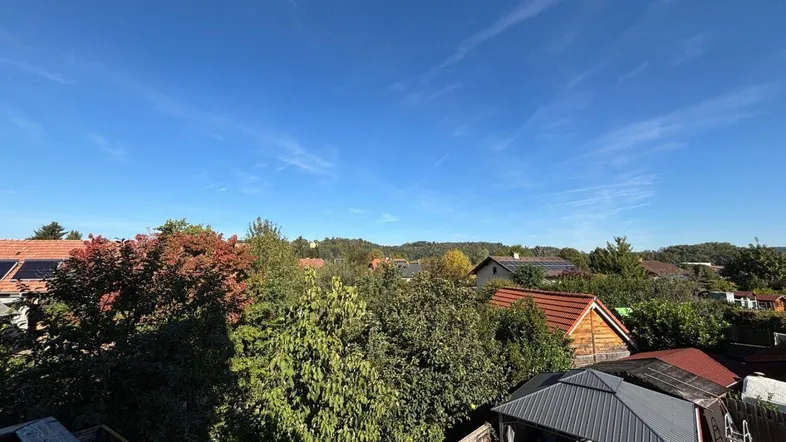 Weitblick über Dächer, grüne Bäume und entfernte Hügel unter einem klaren blauen Himmel.