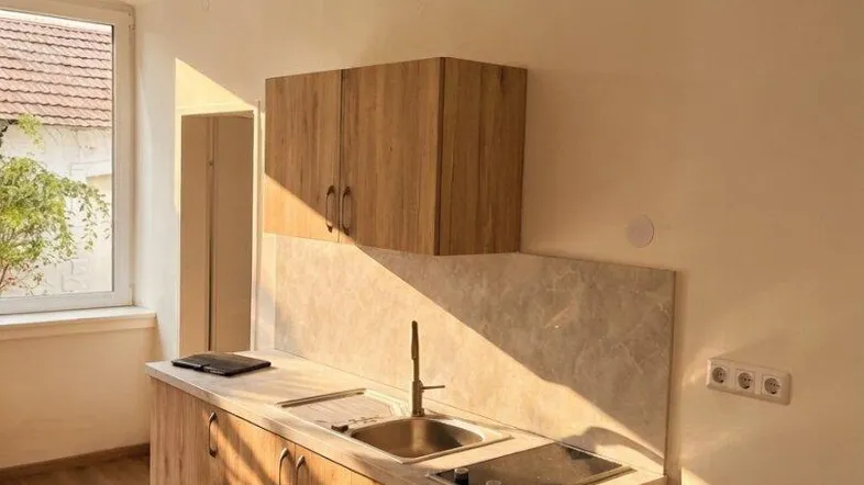 Kleine Küchenzeile mit Holzschränken und Spüle, beleuchtet von natürlichem Sonnenlicht durch das Fenster.