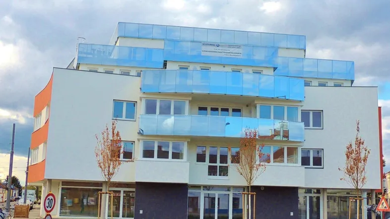 Modernes Mehrfamilienhaus mit weißer Fassade, orangefarbenen Akzenten und blauen Glasbalkonen unter bewölktem Himmel.