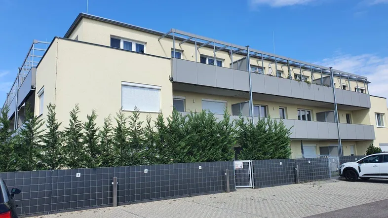 Modernes Mehrfamilienhaus mit Balkonen, umgeben von gepflegten Hecken und Parkplätzen unter blauem Himmel.