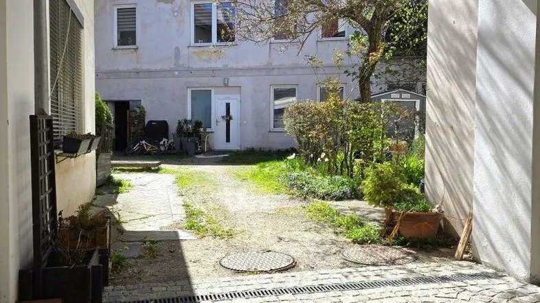 Blick durch einen Torbogen auf einen gepflasterten Innenhof mit Grünflächen und einem Gebäude im Hintergrund.