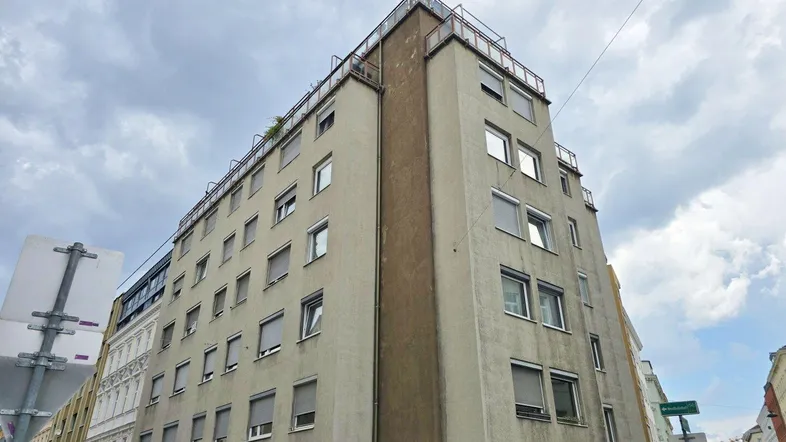 Gebäudeansicht von der Straßenecke mit mehreren Stockwerken und vielen Fenstern unter bewölktem Himmel.