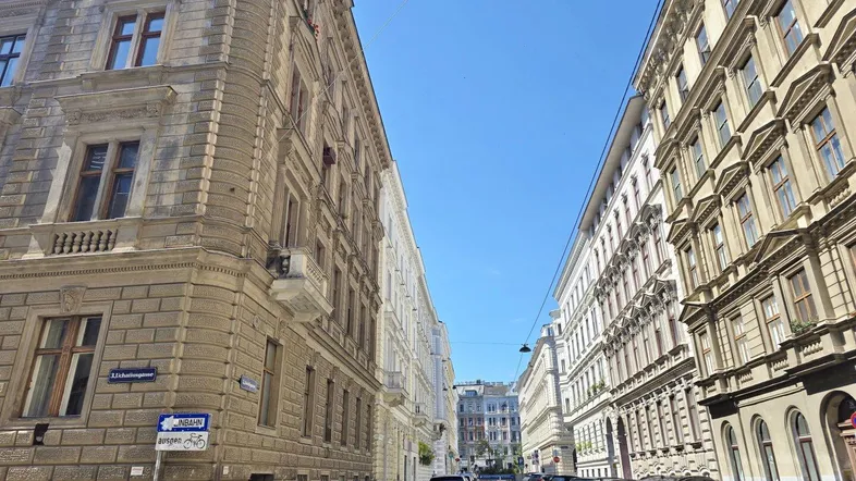 Historische Gebäude säumen eine sonnige Straße in der Stadt, mit geparkten Autos und klarem Himmel.
